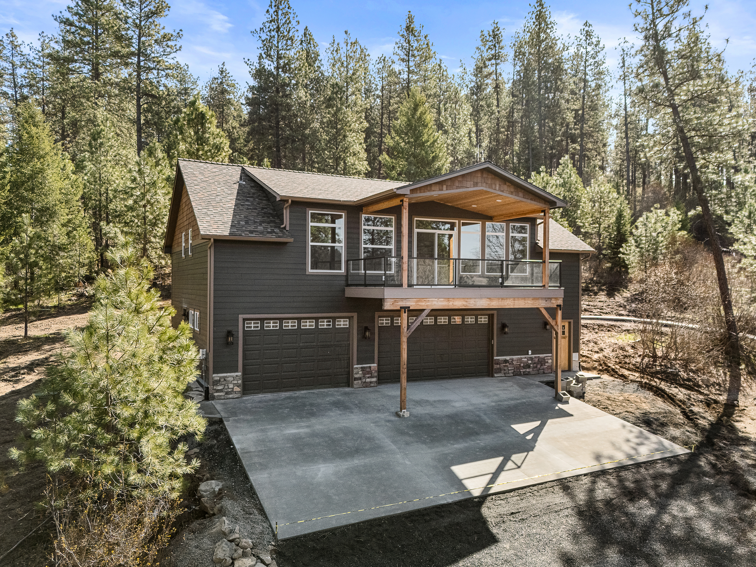 Custom home with dark siding, covered balcony, three-car garage, and concrete driveway surrounded by pine trees in Spokane.