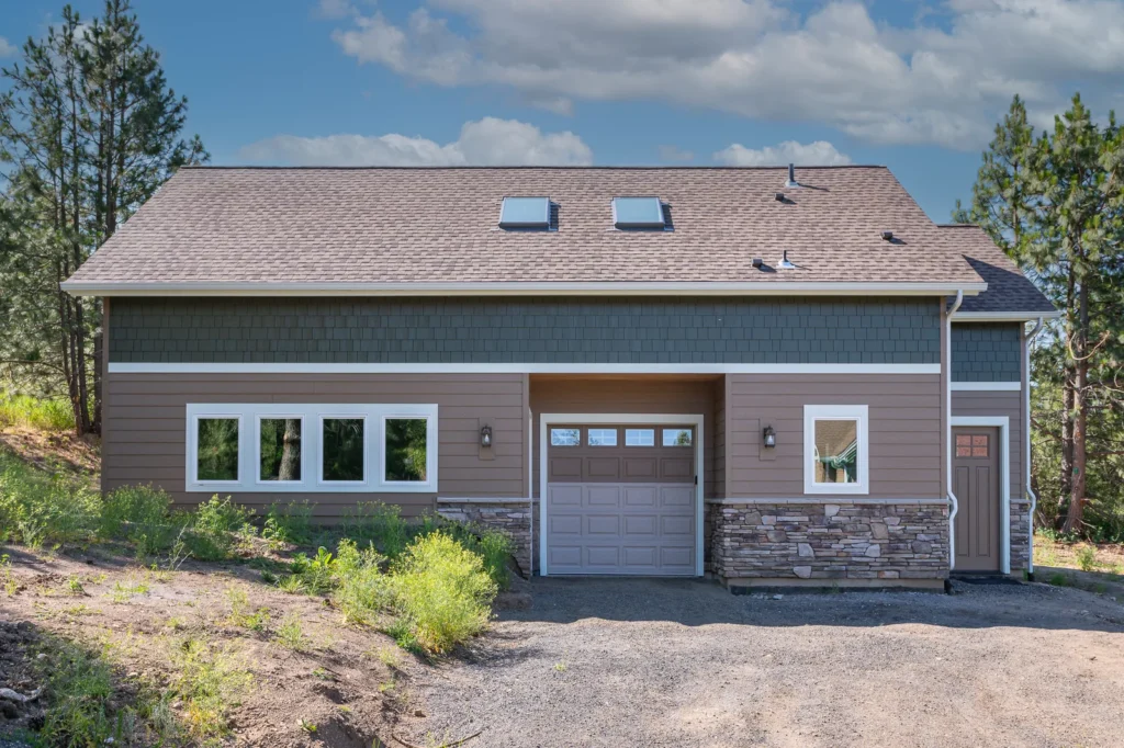 Custom garage exterior with tan siding, stone veneer, overhead garage door, skylights, and wooded Spokane-area setting.
